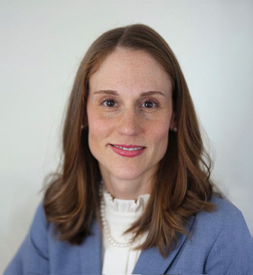 A woman with red hair wearing a light blue blazer and against a soft gray background.