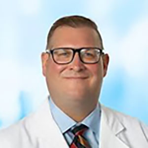 A man with short cut hair, wears a white lab coat and red tie against a blue cloud like background.