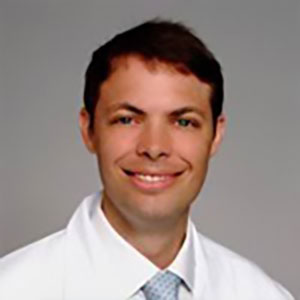 A younger male doctor with scruffy dark hair wears a white lab coat, and tie, smiles in front of a smooth, solid gray background.