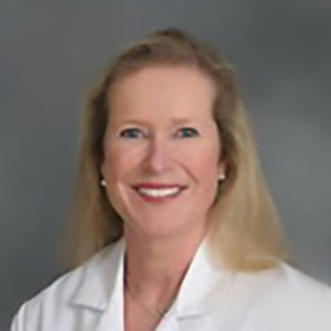 A light haired woman with bright teeth and white lab coat smiles in front of a smooth gray backdrop.